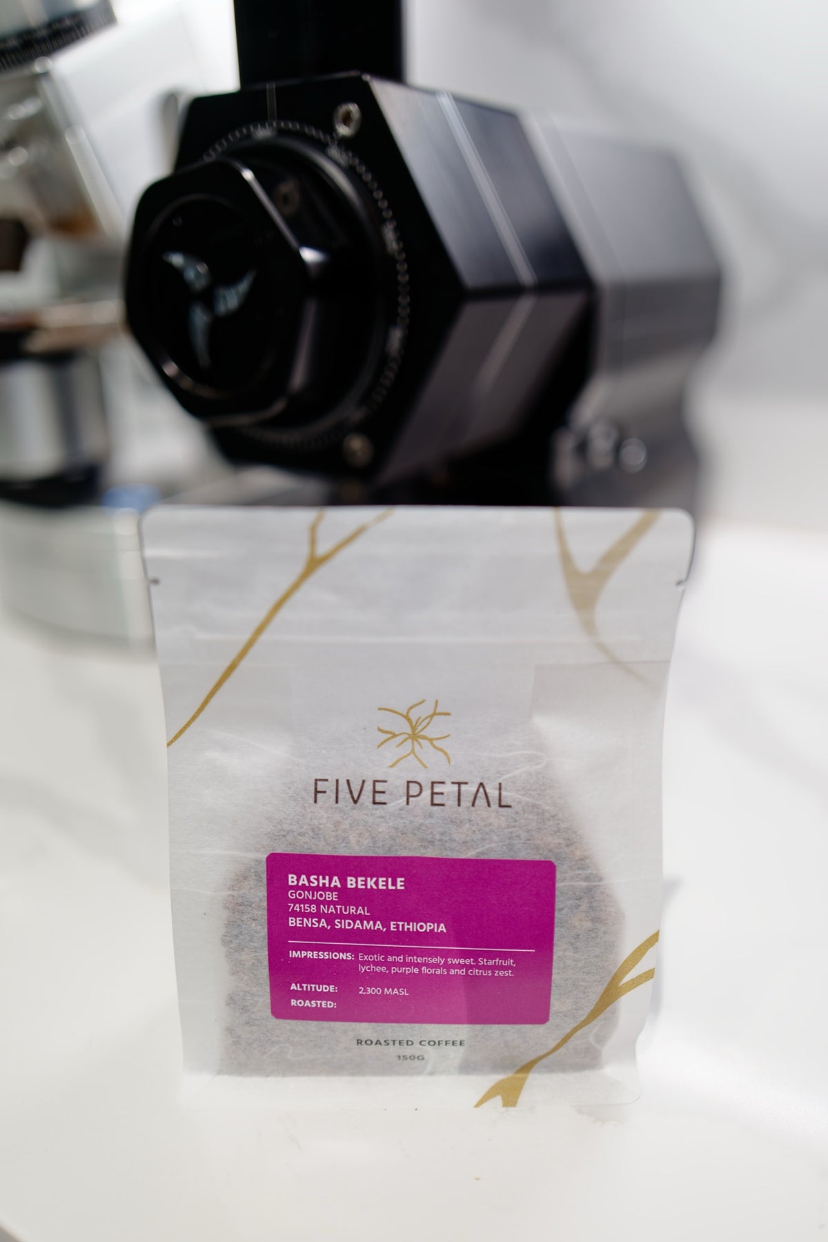150g bag of Basha Bekele Gonjobe Natural coffee standing in front of a black coffee grinder.